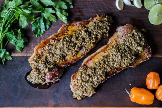 Title: "Chimichurri-Infused Grilled Delicacy" Alternative Text: Two succulent grilled meat strips generously coated with vibrant chimichurri sauce, placed on a dark wooden background accompanied by fresh parsley leaves.